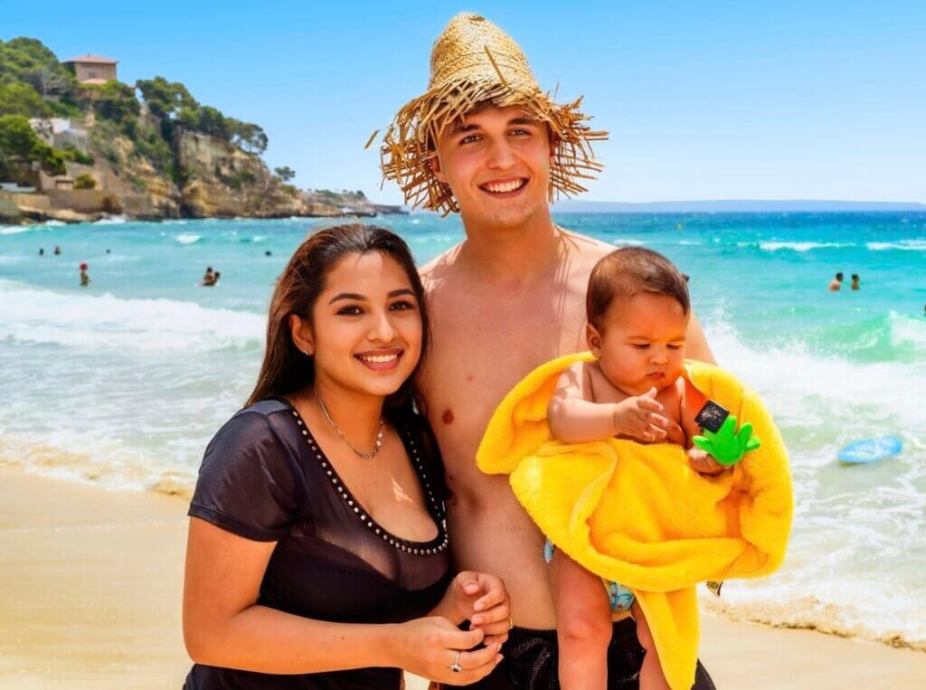 Une famille souriante avec un bébé sur une plage ensoleillée avec la mer en arrière-plan.