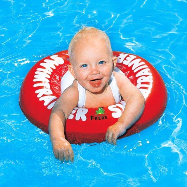 Bébé blond souriant nageant avec une bouée Swimtrainer classique rouge.